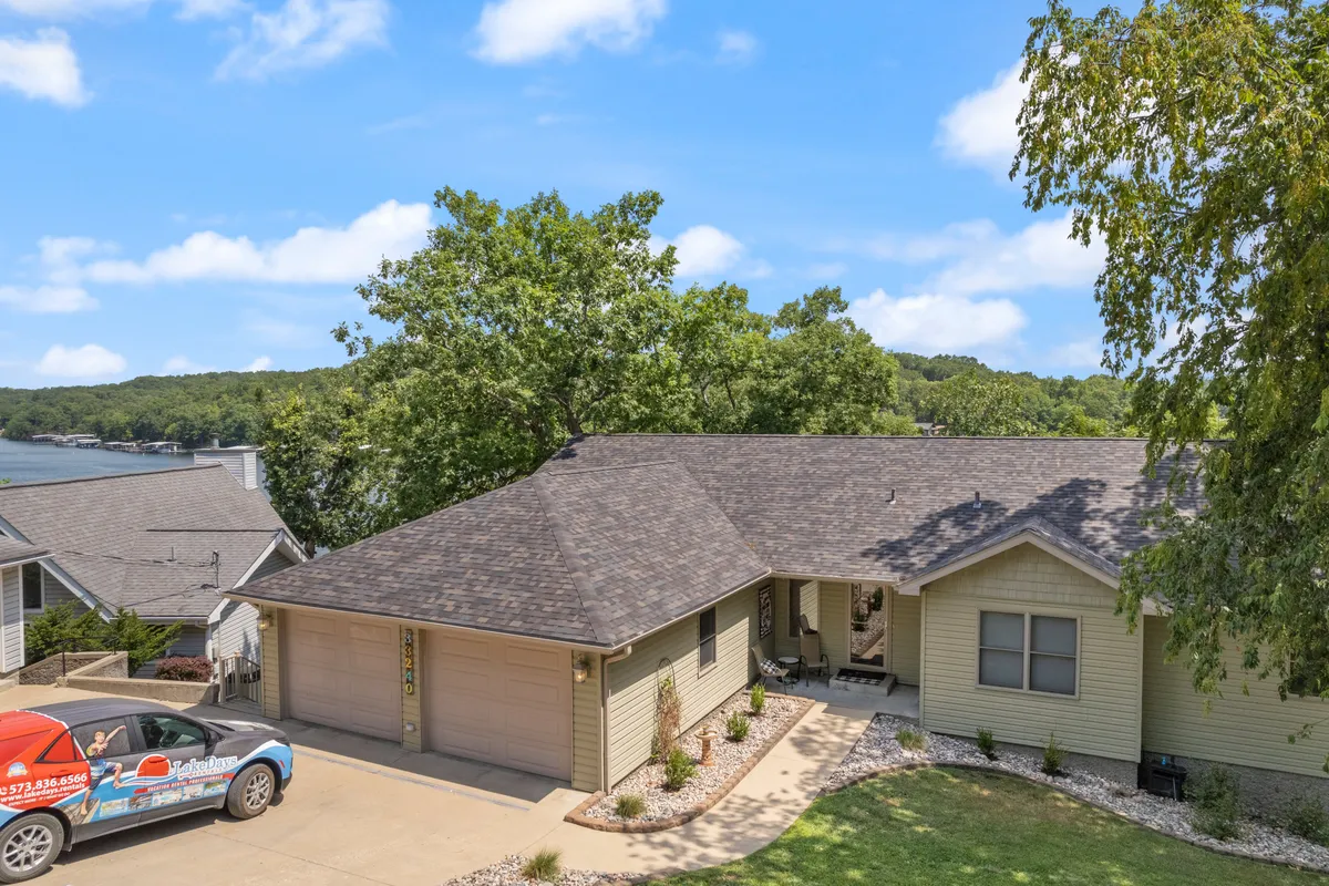 A wide view of the house exterior set against lush greenery with water visible in the background. The driveway, front entrance, and landscaped yard highlight the inviting connection between the home and its natural surroundings.