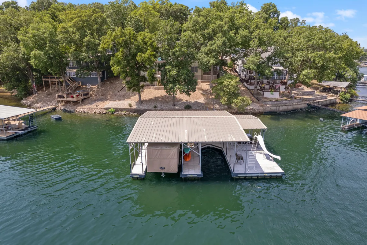 A sunlit dock stretches out onto the lake, complete with a water slide, a nearby boathouse, and lush trees framing the scene. The tranquil water is perfect for swimming, paddling, or launching a kayak. The dock invites guests to enjoy a variety of lakefront adventures, from sunbathing and swimming to evening relaxation by the water.