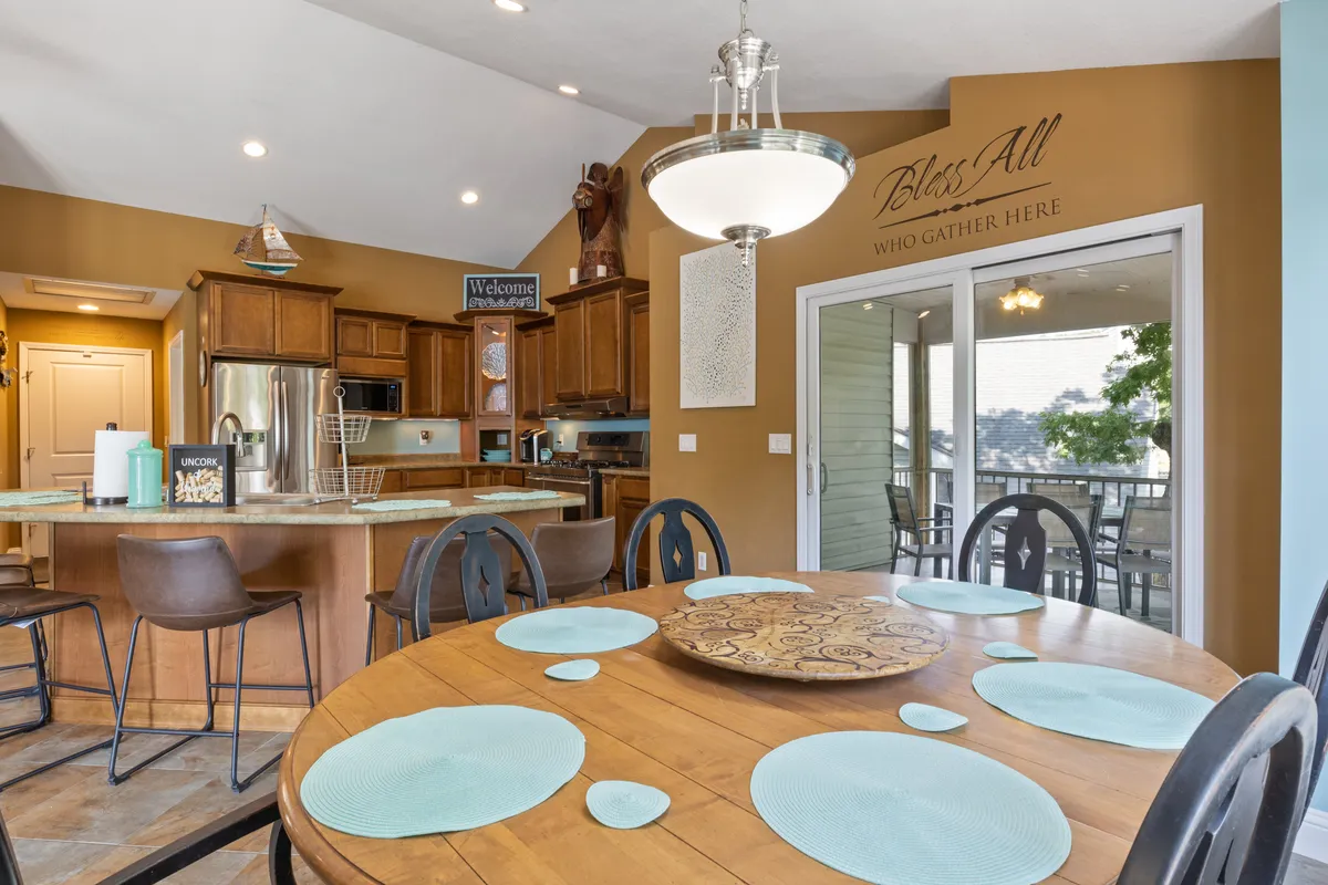 A bright, open-concept interior featuring a spacious dining table adjacent to a kitchen island, all set against a backdrop of large windows that overlook the picturesque lake. The scene is set for family meals or lively gatherings, with the kitchen and dining area seamlessly connected for entertaining and togetherness.