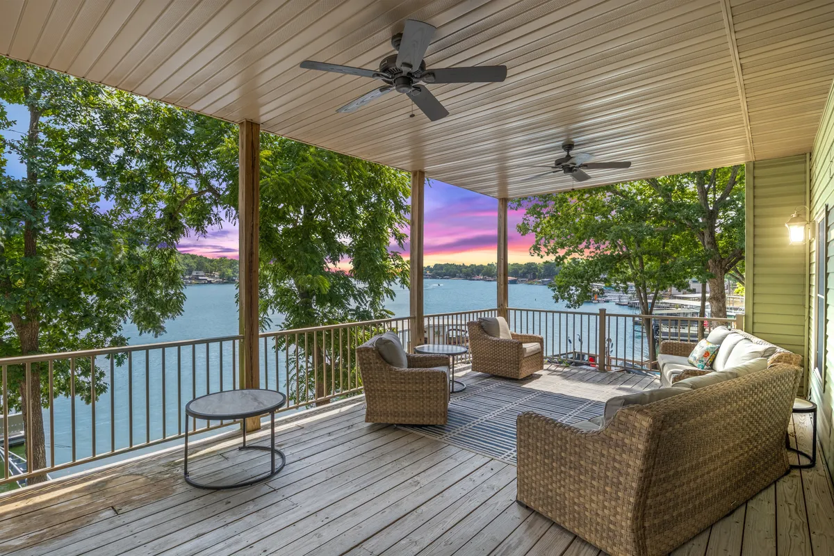 The image showcases an inviting outdoor seating area overlooking a tranquil lake, with a breathtaking, colorful sunset painting the sky. The serene setting is perfect for relaxing, entertaining, or quietly enjoying nature's beauty. The expansive deck seamlessly connects to the natural landscape, offering panoramic water views and a peaceful atmosphere.