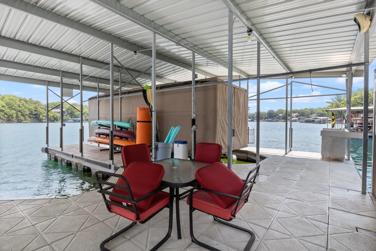 Red chairs and a circular table are arranged on a spacious dock, with kayaks nearby and a sweeping water view. The scene evokes easy access to water activities and relaxed outdoor dining.
