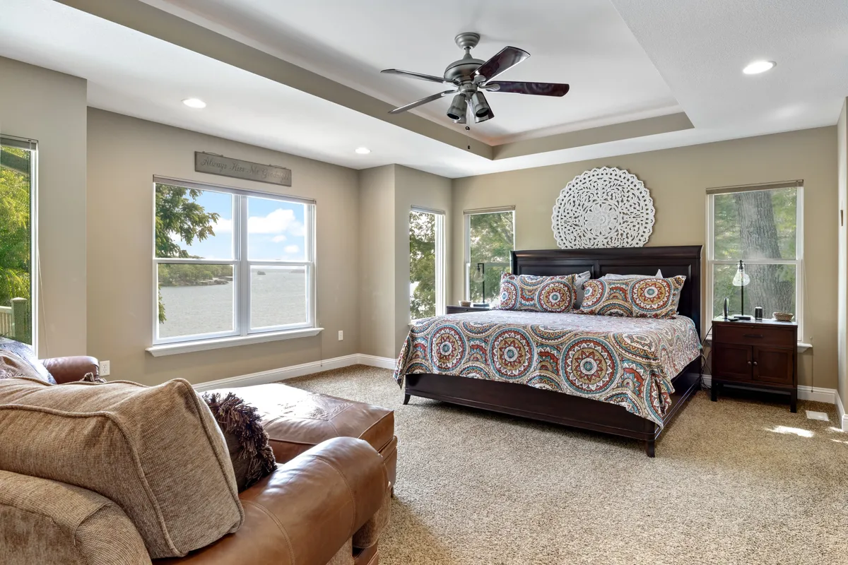 A cozy bedroom featuring a bed with colorful bedding positioned to face expansive windows. Natural light fills the room, and a comfortable seating area is placed near the window, with decorative wall art adding personality. The windows offer tranquil lake views, enhancing the room's peaceful mood.